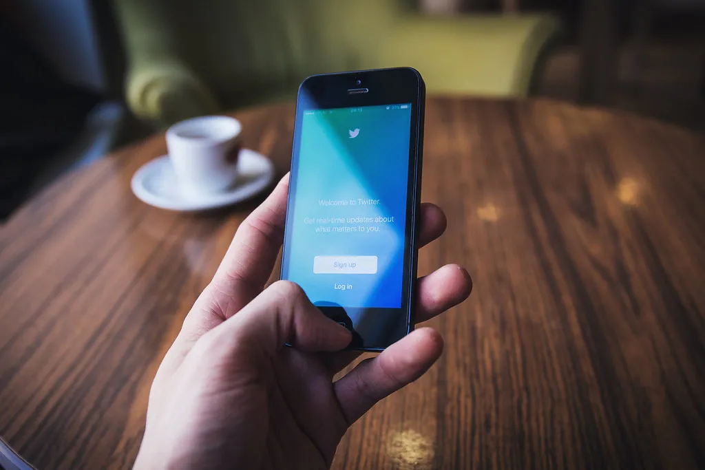 A hand holding a smartphone opening a social media app beside a cup of coffee