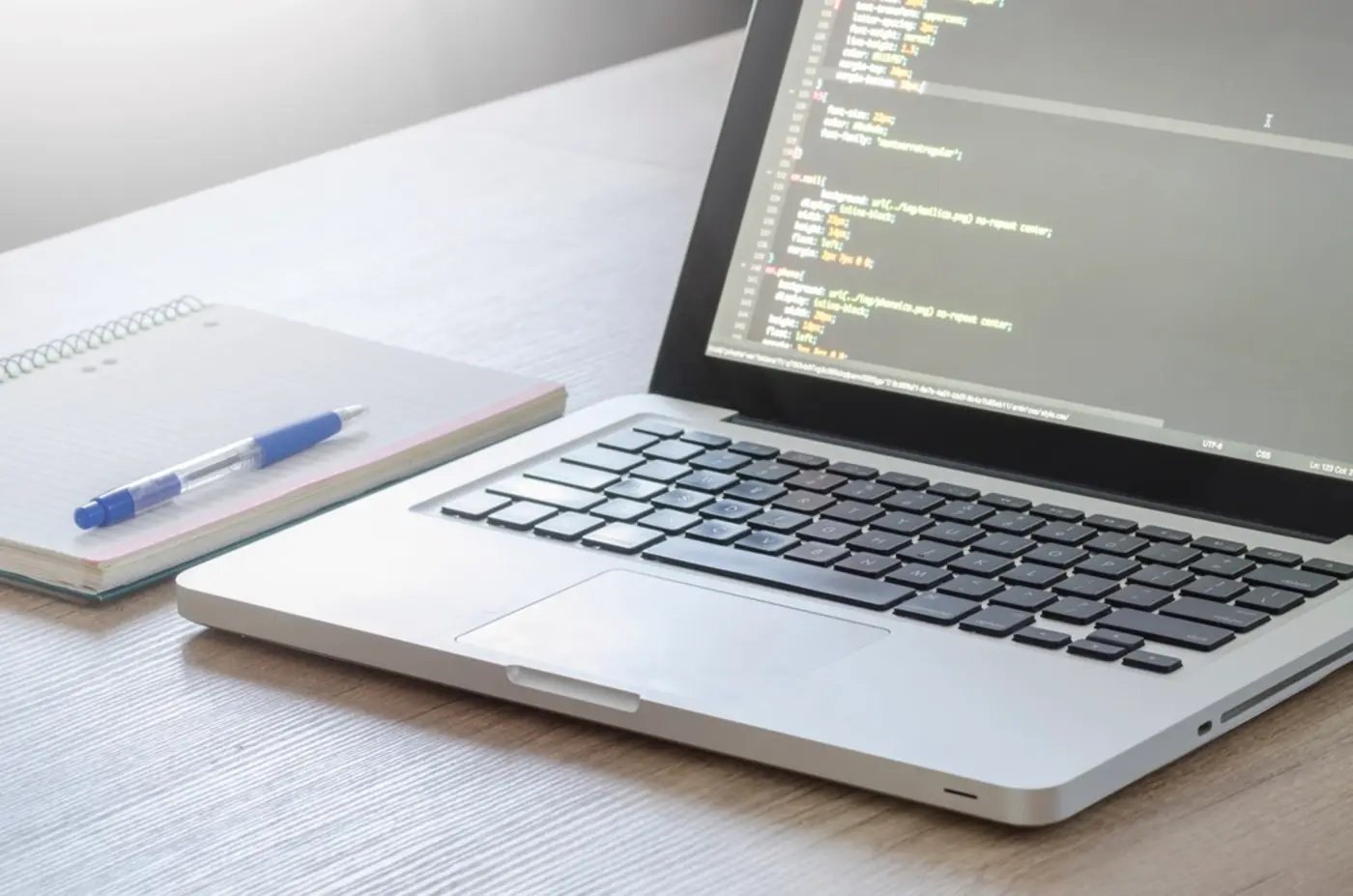 A developer's desk with a laptop showing hand-written website code next to a notepad